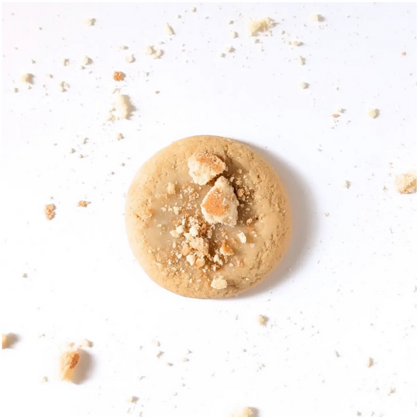 A round golden-brown cookie topped with crumbled biscuit pieces, centered on a white background with scattered crumbs around it.