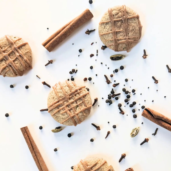 Three round spiced chai cookies with brown icing drizzle are arranged on a white background with cinnamon sticks, cloves, and cardamom seeds as styled props.