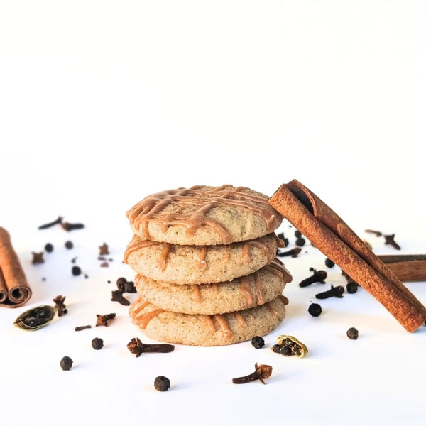 A stack of four spiced chai cookies with a drizzle of icing, styled with cinnamon sticks, cloves, and nutmeg on a white background.