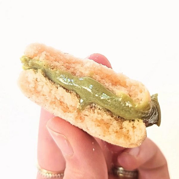 A hand holding a broken strawberry matcha cookie with the matcha filling oozing out of the center on a white background
