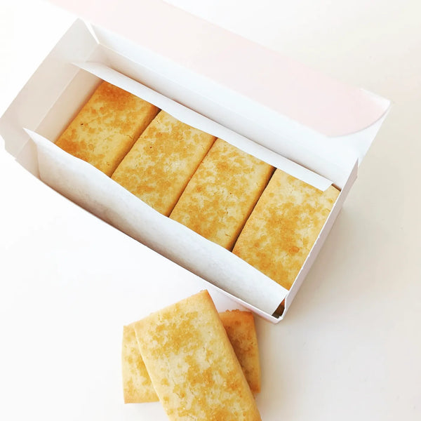 Four rectangular shortbread cookies topped with coarse sugar crystals arranged in a light pink paper box, with two additional cookies placed below it.|