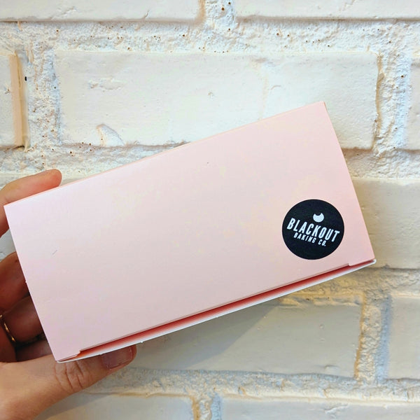 A light pink paper box in front of a white brick background with a Blackout Baking Co. sticker on the corner.